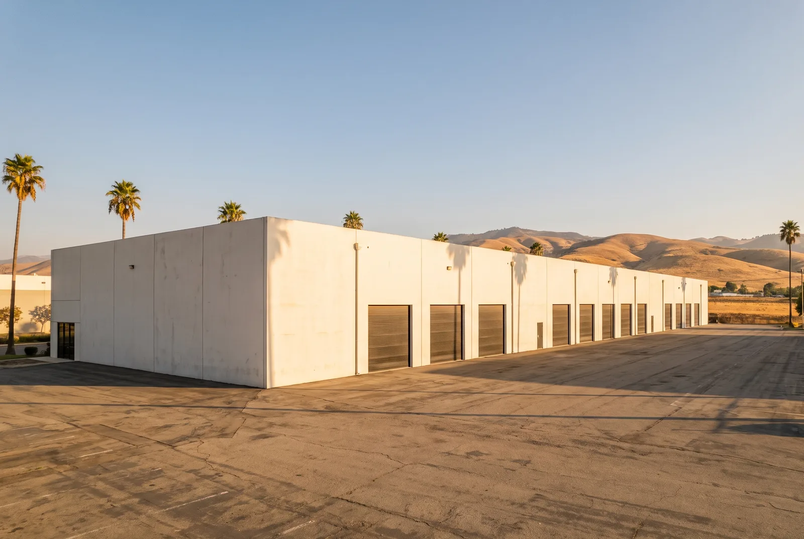 Exterior of a single-story concrete tilt-up warehouse with a row of closed dock doors and an empty truck court, with palm trees and dry golden hills behind it