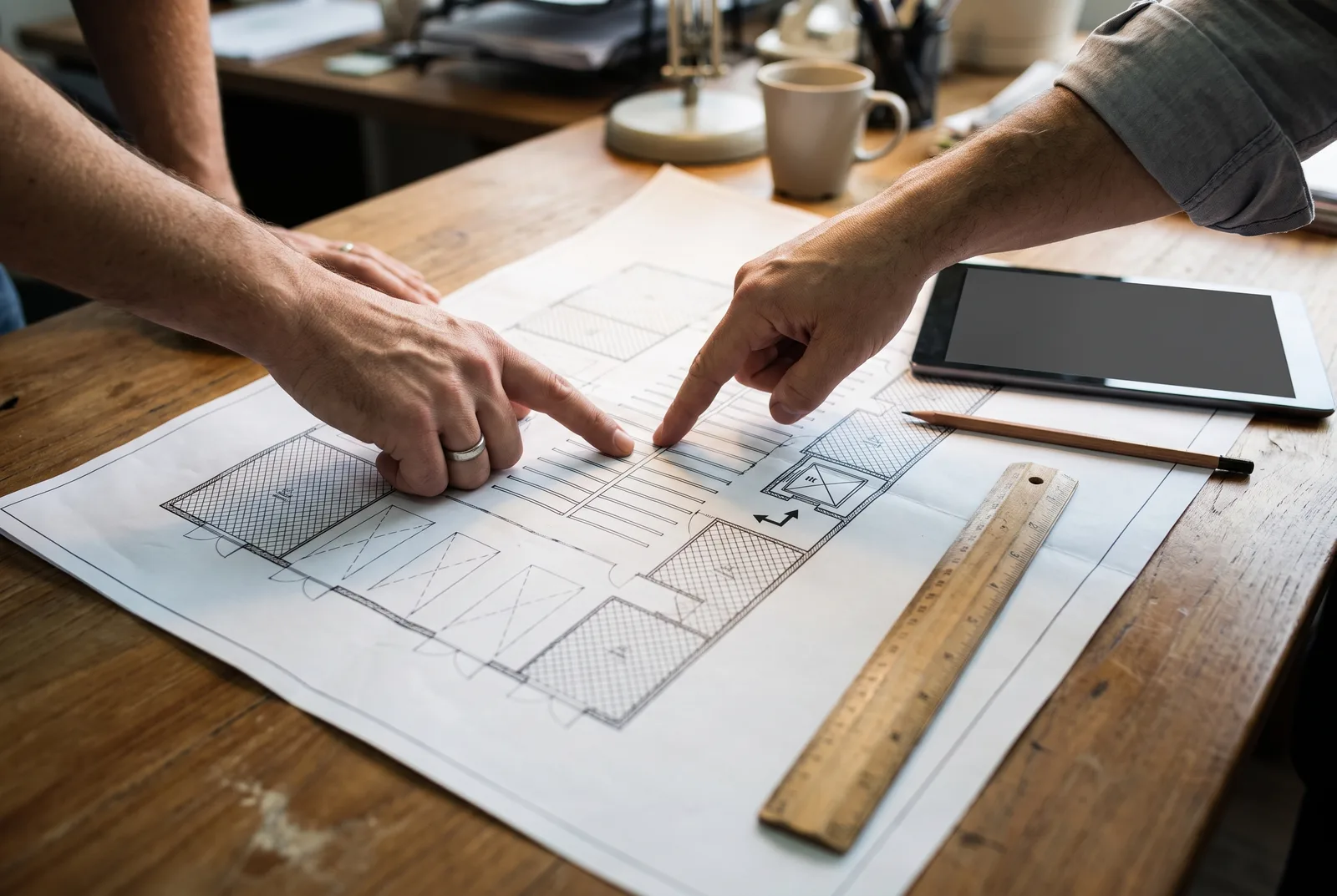 Two people's hands pointing at a printed architectural floor plan of a mezzanine layout on a wooden desk, with a wooden ruler, a pencil and a tablet nearby