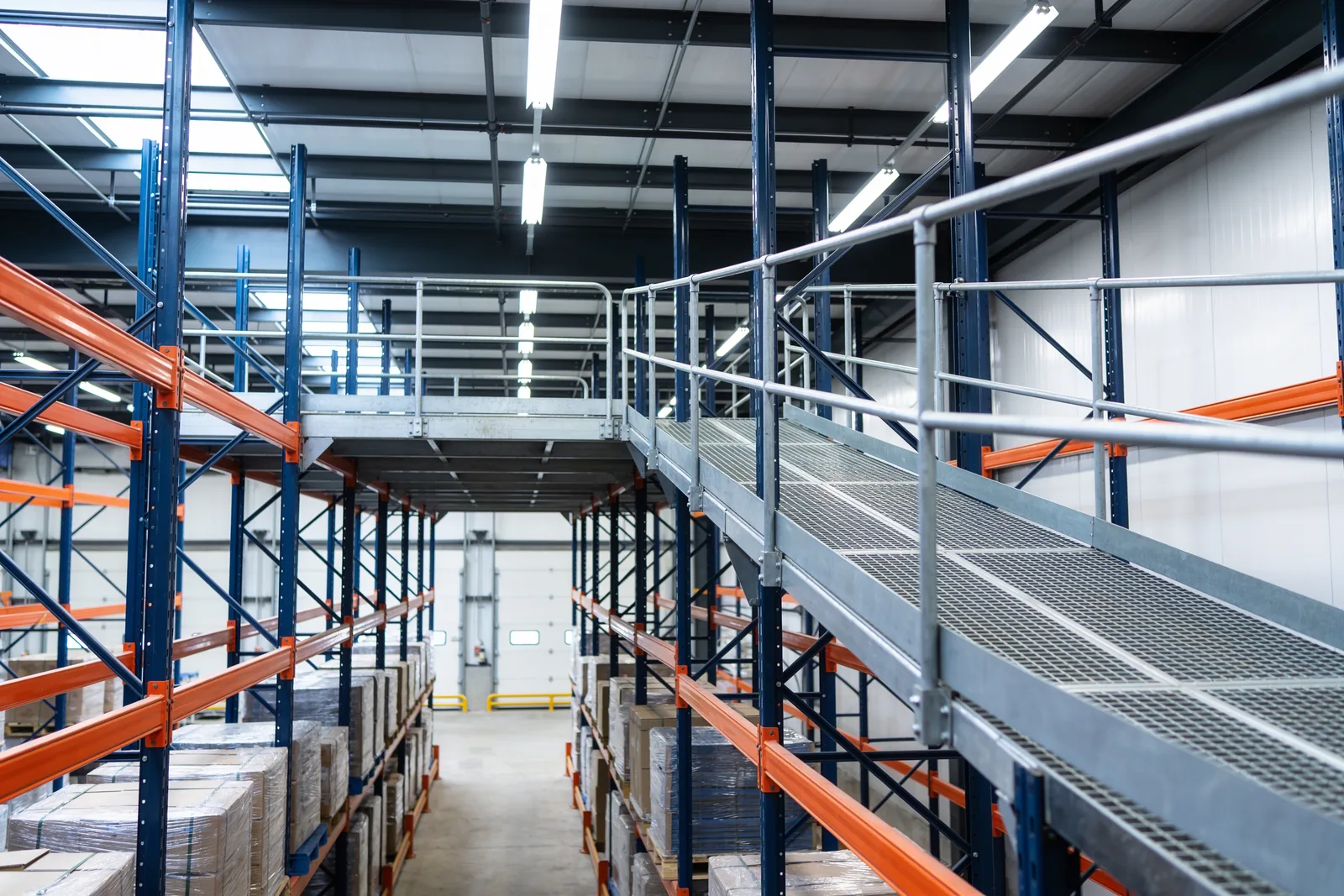 Elevated steel walkway with a bar-grating deck and galvanized guardrails running above blue-grey pallet racking in a warehouse, with a stair rising to the platform and loaded pallets on orange beams below