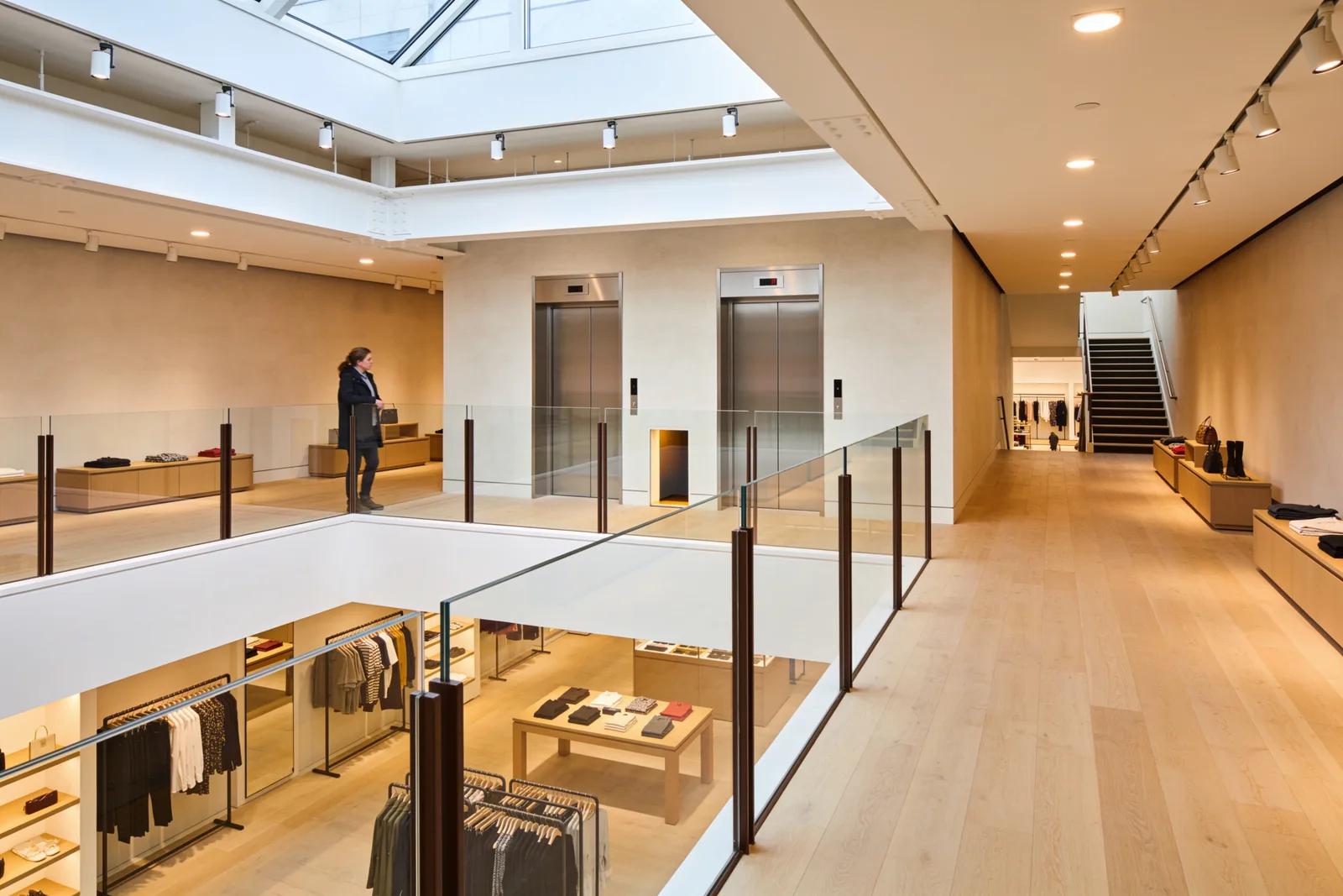 View from a retail mezzanine level: a clear-glass guardrail in slim bronze posts runs along the open edge above the ground-floor sales area, two brushed steel passenger elevator doors face the deck across a plaster wall, low merchandise displays line the pale oak floor, and a second staircase descends at the far end