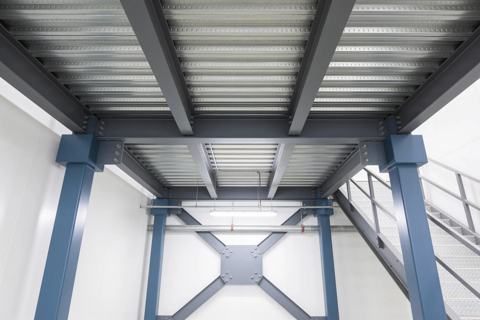 View from beneath a steel mezzanine deck showing grey C-section beams and joists under a corrugated steel deck, slate-blue square columns, a large X-brace with a bolted central gusset between two columns, sprinkler piping, and a galvanized stair at the right