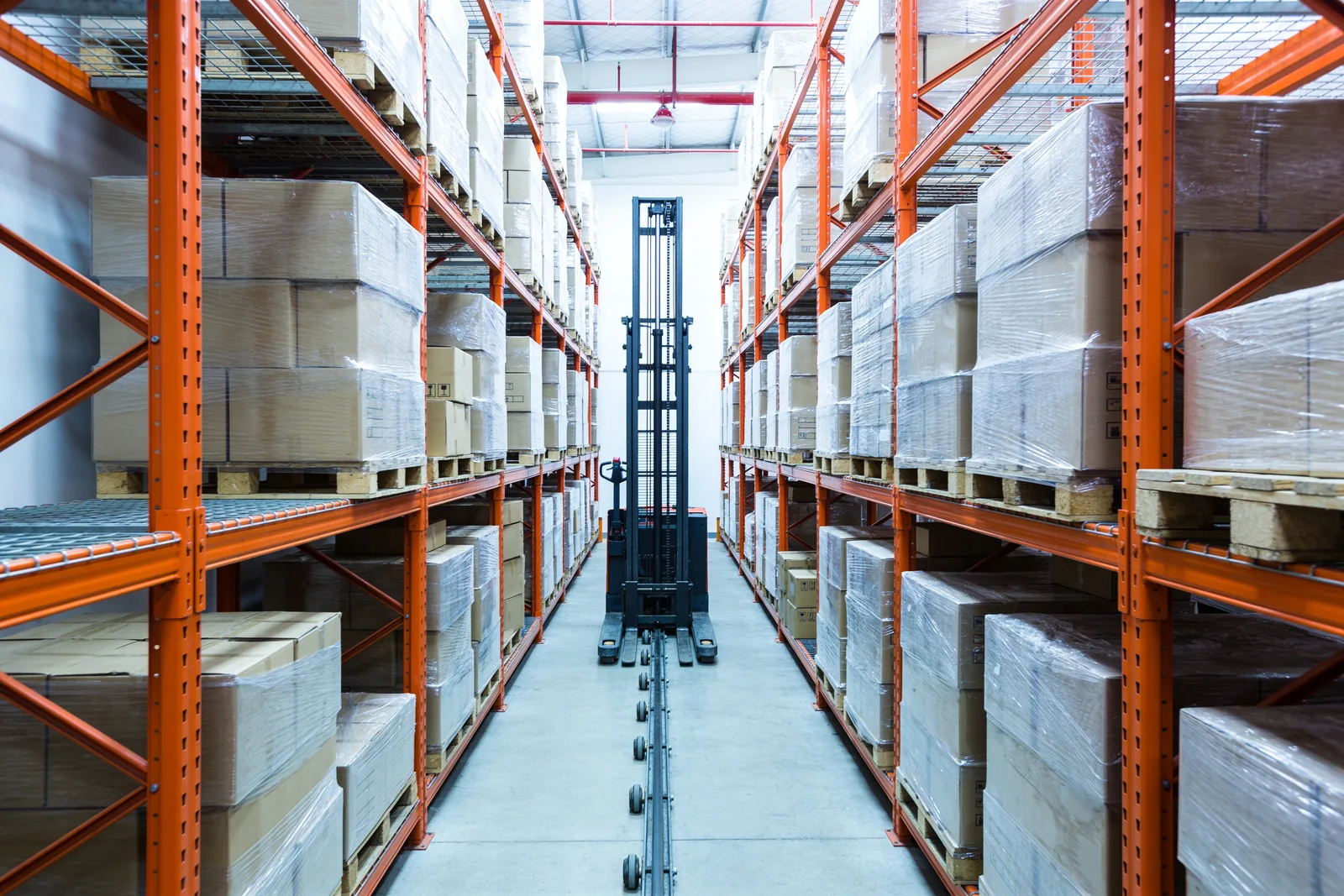 Very narrow aisle between two tall runs of orange pallet racking packed with shrink-wrapped pallets and cartons, with an unoccupied narrow-aisle turret truck with a tall vertical mast standing in the aisle on a guide rail set into the concrete floor, wire mesh decking visible on the beam levels
